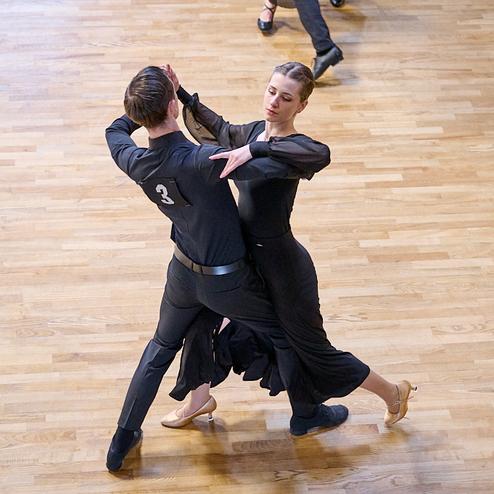 Tanzpaar in schwarzer Kleidung führt einen Paartanz auf einem hellen Holzboden aus.