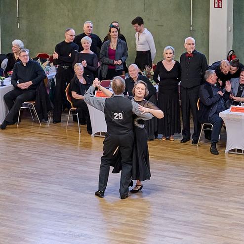 Tanzpaar in eleganter Kleidung während eines Wettbewerbs, umgeben von zuschauenden Gästen an festlich gedeckten Tischen.