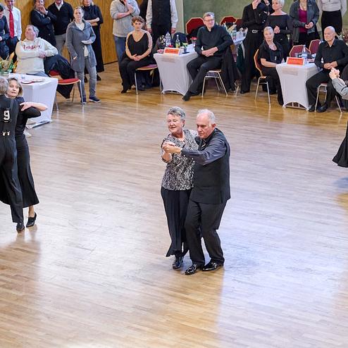 Tanzpaare bei einer Veranstaltung in einem großen Saal, Zuschauer sitzen an Tischen und beobachten das Geschehen.