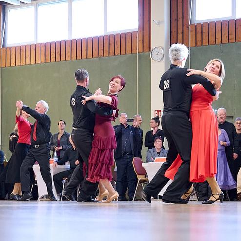 Tanzende Paare in eleganter Kleidung auf einer Bühne, Zuschauer sitzen an Tischen im Hintergrund.