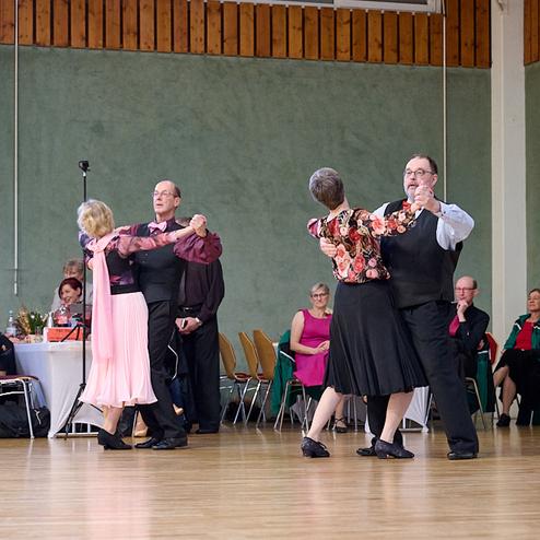 Paare tanzen bei einer Tanzveranstaltung, Zuschauer beobachten und applaudieren im hinteren Bereich des Saals.
