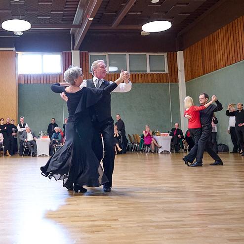 Tanzpaar wirbelt in eleganter Kleidung über eine Tanzfläche, Zuschauer applaudieren im Hintergrund.