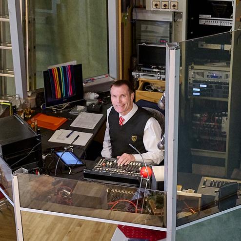 Mann in einem Kontrollraum sitzt an einem Mischpult, umgeben von Technik und Bildschirmen, lächelnd in die Kamera.