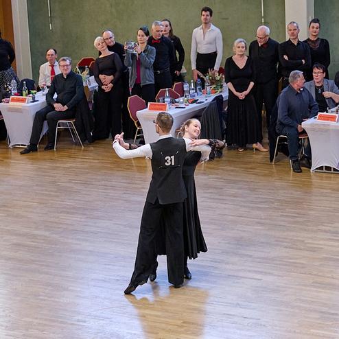 Tanzendes Paar in eleganter Kleidung bei einem Wettkampf, umgeben von zuschauenden Gästen in festlicher Stimmung.