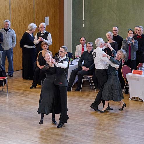 Tanzpaare treten bei einem Wettbewerb in einem festlich dekorierten Saal auf, während Zuschauer applaudieren.