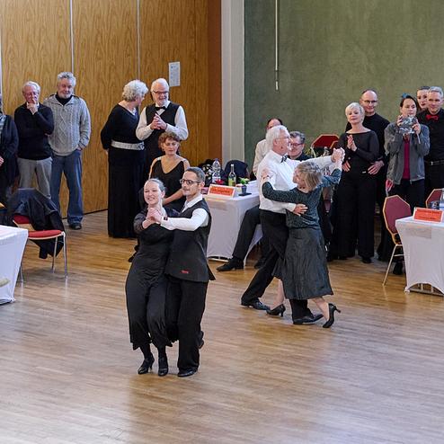 Tanzende Paare in eleganter Kleidung umgeben von zuschauenden Gästen in einem festlichen Saal.
