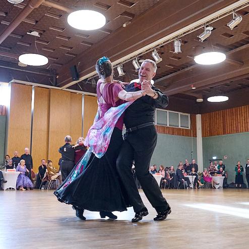 Tanzpaar in eleganter Kleidung tanzt auf Parkettboden vor zuschauendem Publikum in festlicher Atmosphäre.