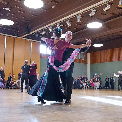 Tanzpaar in eleganter Kleidung tanzt in einem festlichen Saal vor Publikum, umgeben von Tischen und Stühlen.