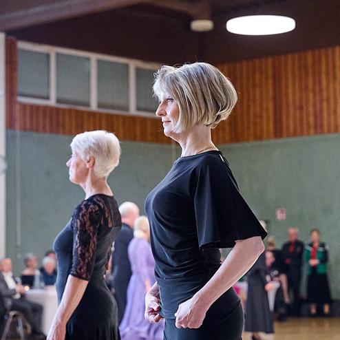 Zwei Frauen stehen in eleganter Kleidung in einer Tanzposition auf einer Turnierbühne, Zuschauer im Hintergrund.