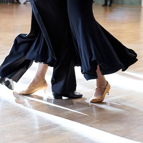 Tanzende Füße in eleganter Kleidung auf einem Holzparkettboden, mit Lichtstrahlen im Hintergrund.