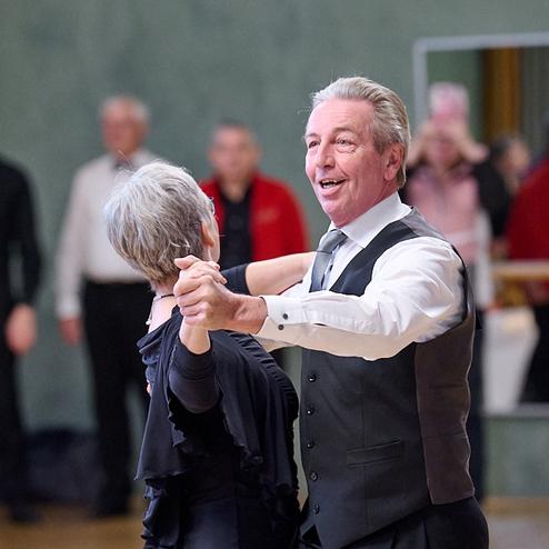 Tanzendes Paar in eleganter Kleidung, umgeben von Zuschauern in einem festlichen Veranstaltungsraum.