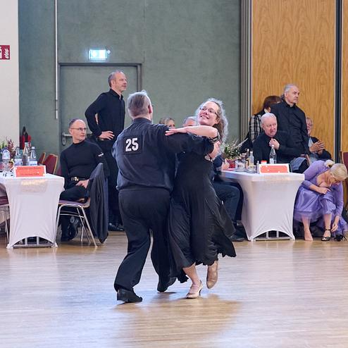 Tanzpaar in eleganter Kleidung tanzt in einem festlichen Saal, während Zuschauer an Tischen beobachten.