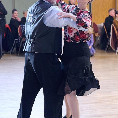 Tanzpaar in eleganter Kleidung beim Tanzen auf einer Tanzfläche, umgeben von weiteren Gästen im Hintergrund.