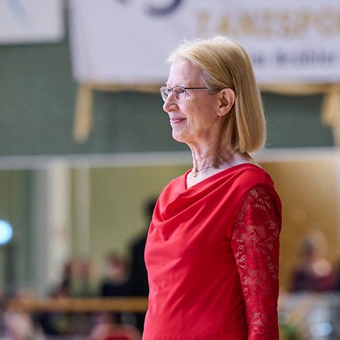 Ältere Dame in rotem Kleid steht lächelnd auf einer Tanzfläche während einer Tanzveranstaltung.