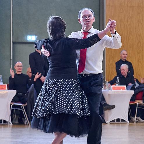 Tanzendes Paar in eleganter Kleidung bei einer Tanzveranstaltung, im Hintergrund applaudierendes Publikum.