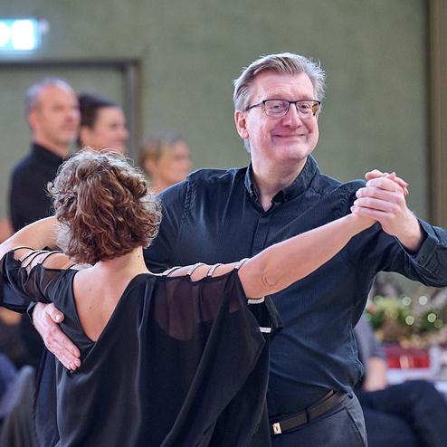 Tanzpaar in formeller Kleidung tanzt lächelnd in einem festlich dekorierten Saal. Menschen im Hintergrund beobachten das Geschehen.