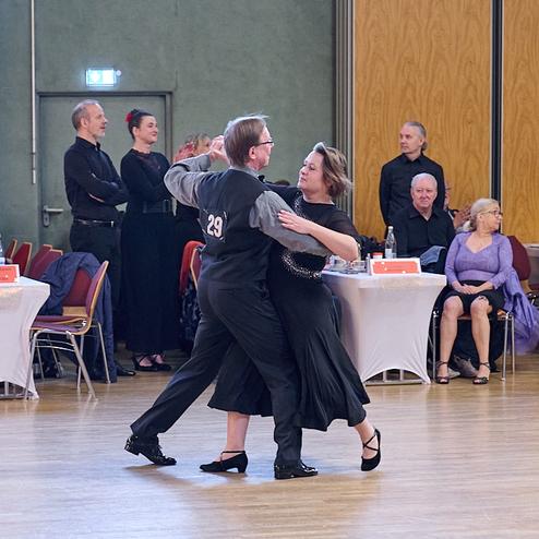 Tanzendes Paar in eleganter Kleidung auf einer Tanzfläche, umgeben von Zuschauern und Tischen.