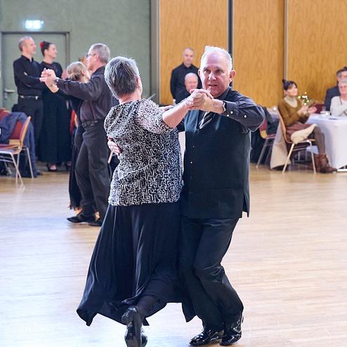 Tanzpaar in eleganter Kleidung tanzt auf einer Holzfläche, während Zuschauer an Tischen applaudieren.