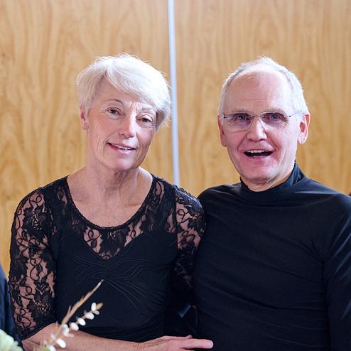 Ein Paar sitzt nebeneinander und lächelt in die Kamera, vor einem hellen Holzhintergrund.