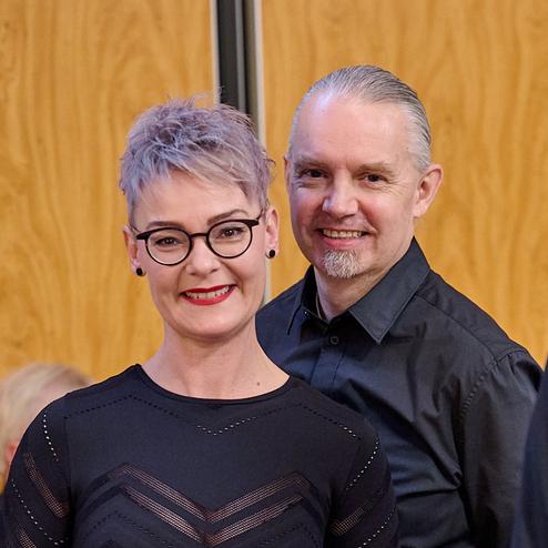 Lächelndes Paar in formeller Kleidung vor einer Holzvertäfelung, im Hintergrund sind weitere Personen unscharf zu sehen.