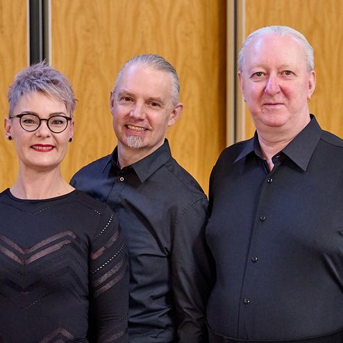 Drei Personen in schwarzer Kleidung stehen lächelnd vor einer Holzpaneelwand, mit einer Person im Hintergrund.