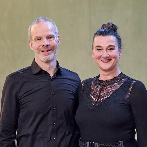 Paar in formeller Kleidung lächelt in einem Raum mit grünem Hintergrund, im Hintergrund sind Tische und Dekoration sichtbar.