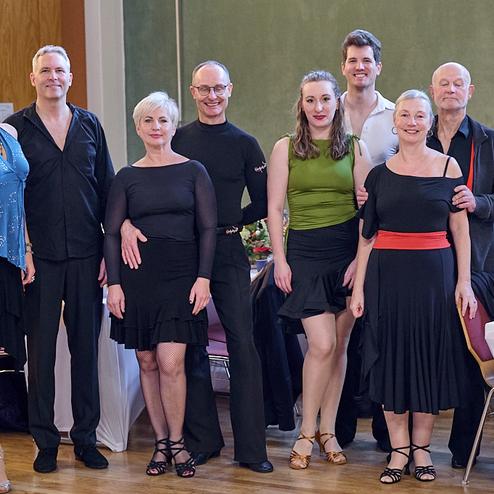 Gruppe von acht Tänzern in eleganter Kleidung steht lächelnd in einem festlichen Raum. Tisch mit Dekoration im Hintergrund.