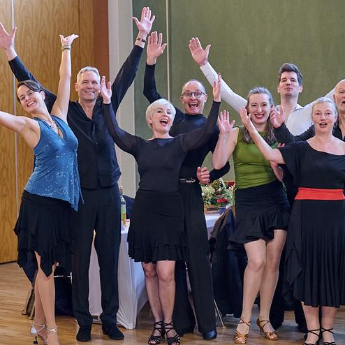 Eine Gruppe von acht tanzenden Personen in eleganter Kleidung hebt die Arme in die Luft und lächelt fröhlich.