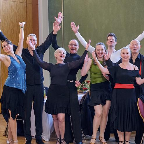Gruppe von tanzenden Menschen in Schwingkleidung hebt die Arme vor einem festlich gedeckten Tisch.