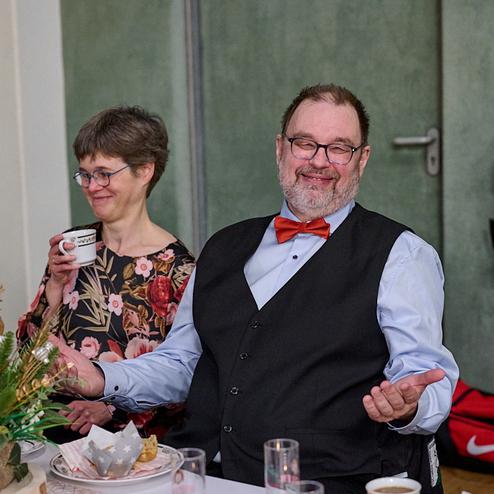 Lächelnder Mann mit Brille und Fliege sitzt an einem Tisch, während eine Frau mit Kaffeetasse neben ihm steht.