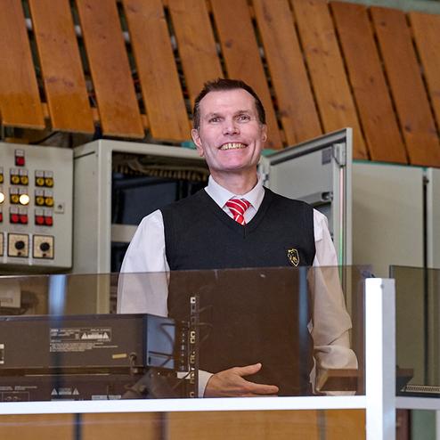 Lächelnder Mann in Weste hinter einem Empfangstresen mit Technik und Steuerungselementen im Hintergrund.