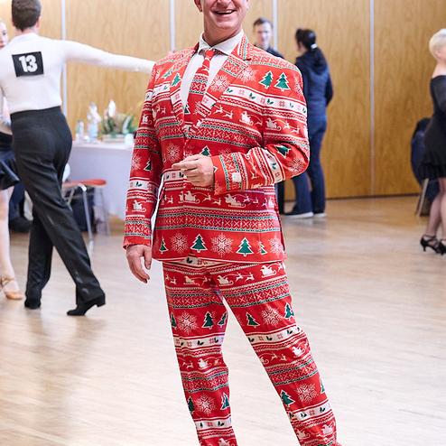 Mann in einem weihnachtlich gemusterten Anzug lächelt auf einem Tanzparkett, im Hintergrund Tänzer und festliche Dekoration.