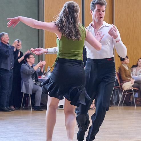 Tanzendes Paar in elegantem Outfit auf einer Tanzfläche, umgeben von Zuschauern bei einer Veranstaltung.