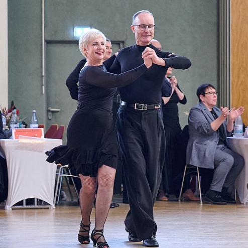 Tanzendes Paar in eleganter Kleidung, umgeben von applaudierendem Publikum in einem Saal.