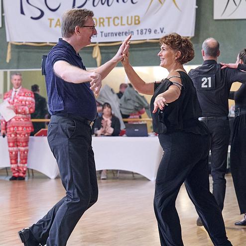 Tanzpaar in formeller Kleidung tanzt fröhlich in einem Sportclub, im Hintergrund Zuschauer und ein Banner des Clubs.