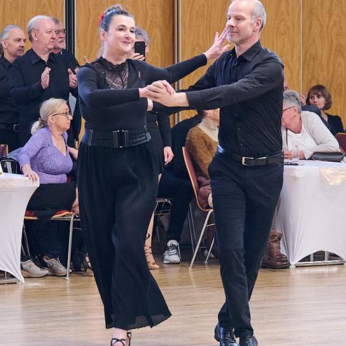 Tanzpaar in eleganter Kleidung beim Tanzen auf einer Veranstaltung, Zuschauer beobachten im Hintergrund.