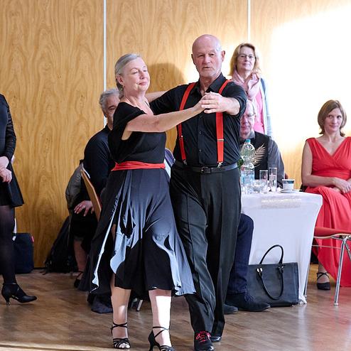 Tanzendes Paar in eleganter Kleidung, umgeben von Zuschauern in festlicher Atmosphäre. Holzfußboden im Hintergrund.