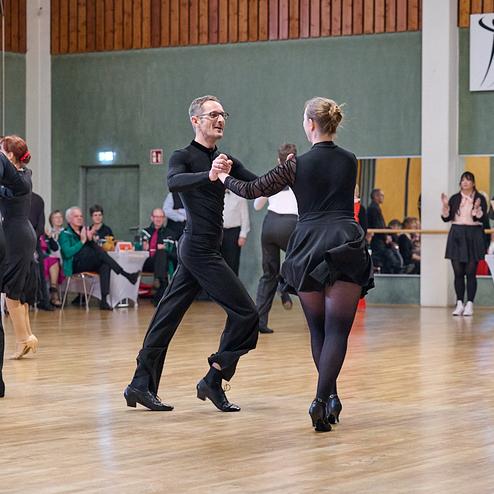 Tanzpaare in eleganter Kleidung bei einem Tanzwettbewerb in einem hellen Saal. Zuschauer applaudieren im Hintergrund.