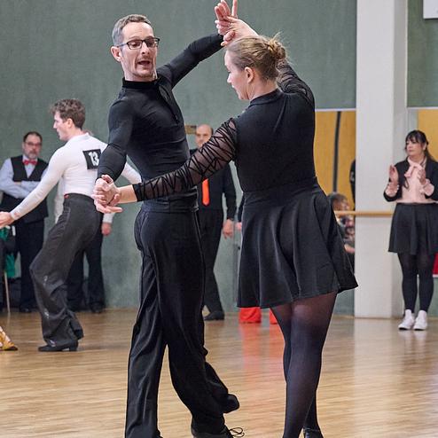 Tanzpaar in eleganter Kleidung, das auf einer Tanzfläche zu einem Wettbewerb tanzt. Zuschauer im Hintergrund applaudieren.