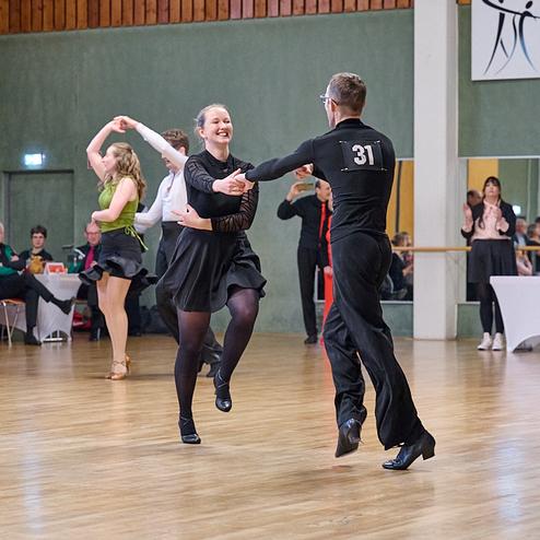 Tanzpaare in eleganter Kleidung beim Wettkampf auf einer Tanzfläche, Zuschauer im Hintergrund applaudieren.
