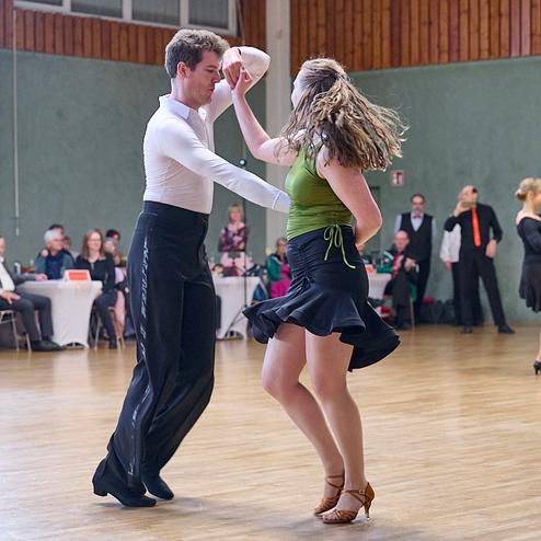 Tanzpaar in eleganter Tanzhaltung auf einer Tanzfläche, Zuschauer sitzen an Tischen im Hintergrund.