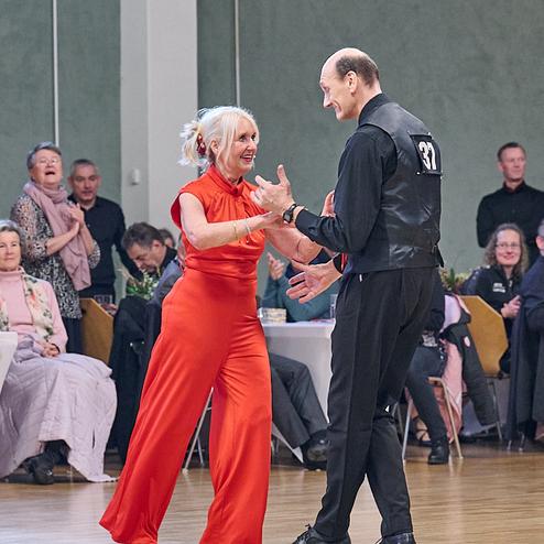 Tanzpaar in eleganter Kleidung steht auf der Tanzfläche und lächelt sich an, im Hintergrund Zuschauer an Tischen.
