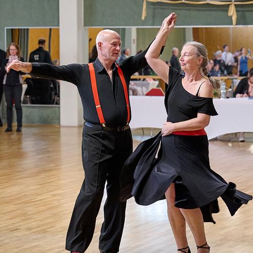 Tanzendes Paar in eleganter Kleidung, der Mann mit roten Hosenträgern und die Frau in einem schwarzen Kleid, auf einer Tanzfläche.