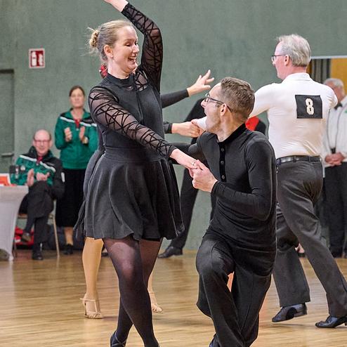 Tanzpaar in formeller Kleidung, während eines Tanzwettbewerbs auf Holzfläche, Publikum im Hintergrund.