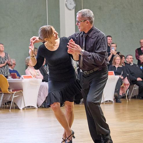 Tanzpaar in eleganter Kleidung während einer Tanzveranstaltung auf einem Holzfußboden, Zuschauer im Hintergrund.