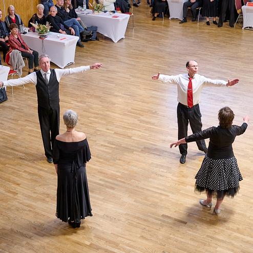 Tanzpaar zeigt eine Choreografie auf einer Holztanzfläche, umgeben von aufmerksam zuschauendem Publikum.
