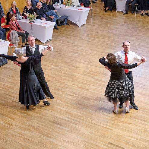 Tanzpaar mit eleganter Kleidung tanzt auf einem Parkettboden, im Hintergrund Zuschauer an Tischen.