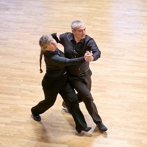Tanzpaar in Schwarz führt eine elegante Tanzbewegung auf einem Holzparkettboden aus.