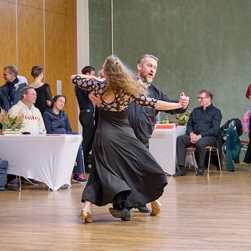 Tanzpaar in eleganter Kleidung führt einen Tango in einem festlichen Saal mit Zuschauern im Hintergrund auf.