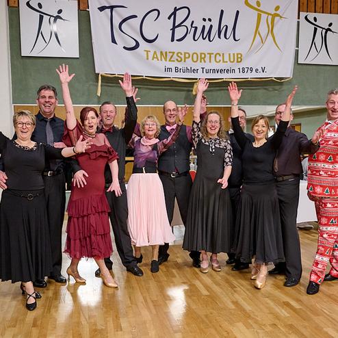 Eine Gruppe von Tänzern in formeller Kleidung posiert fröhlich vor einem Banner des TSC Brühl in einer Tanzhalle.